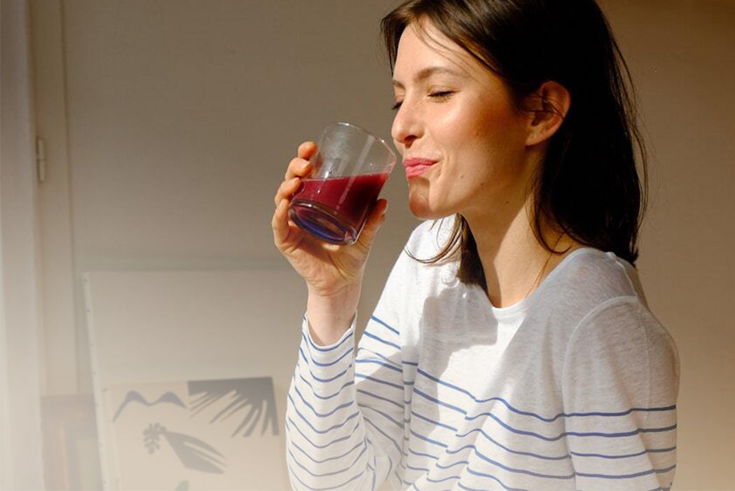 frau dunkle haare augen geschlossen trinkt genussvoll kombucha sitzt im zimmer sonne scheint rein